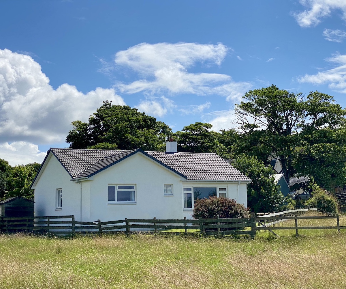 A white bungalow nestled in the lush countryside of Back of Keppoch. The house, with its neat grey roof and white chimneystack, is encircled by a wooden fence and set against a backdrop of mature trees and a vibrant blue sky dotted with fluffy white clouds