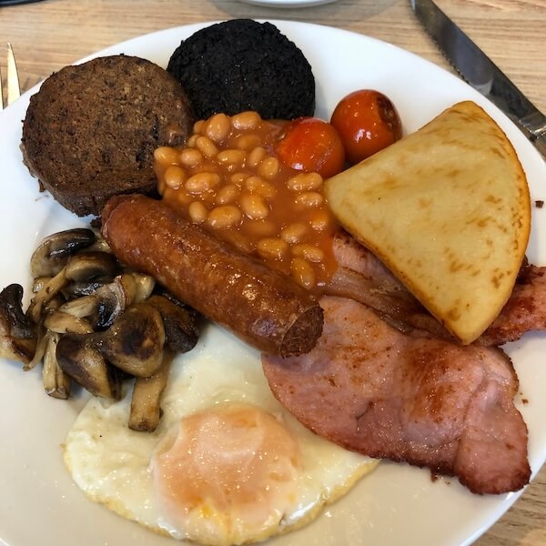 A traditional Scottish breakfast served on a white plate, featuring a fried egg, rashers of back bacon, a sausage, baked beans, grilled tomatoes, sautéed mushrooms, a slice of potato scone, black pudding, and a slice of haggis. The hearty mix showcases classic elements of a full cooked Scottish breakfast.