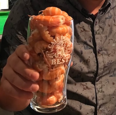A man holds up a Caledonia Best pint glass filled entirely with bright orange langoustine tails, stacked tightly to the brim.
