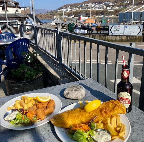Two plates of seafood sit on an outdoor table overlooking a harbor—one with golden battered fish and chips, the other with breaded scampi and chips, both served with salad, tartar sauce, and lemon wedges. A bottle of Fentimans Ginger Beer with a striped straw completes the seaside meal setting.